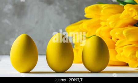 Bouquet de tulipes jaune vif et trois oeufs de Pâques, debout sur une surface en bois blanc sur fond gris, espace de copie Banque D'Images