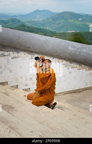 2019-11-06 / Phuket, Thaïlande - UN moine en robe de safran se agenouille pour prendre une photo avec un appareil photo reflex numérique. Banque D'Images