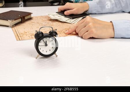 les mains des femmes tiennent un portefeuille en cuir noir avec de l'argent et montrent l'endroit de repos sur la carte du monde. sur une table blanche marron journal et réveil. Banque D'Images