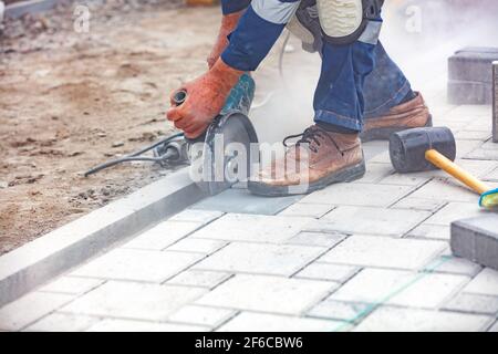 Les mains d'un constructeur à l'aide d'une meuleuse coupent les pavés sur la toile de fond du chantier. Gros plan, arrière-plan flou. Banque D'Images