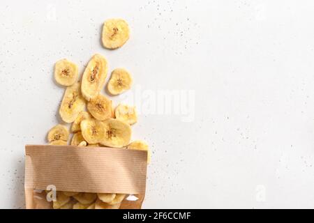 Croustilles de banane de fruits en tranches séchées dans un sac en papier sur fond blanc avec espace de copie. En-cas végétaliens sans sucre. Banque D'Images