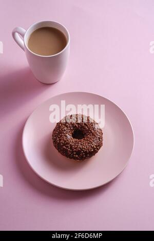 Beignet de chocolat avec saupoudrer sur l'assiette près d'une tasse de café, dessert glacé sucré et boisson chaude sur fond rose minimal, vue en angle Banque D'Images