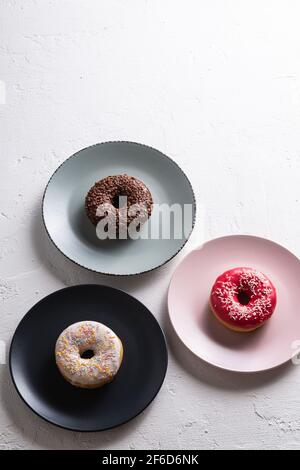 Trois beignets sur des assiettes, chocolat, beignets roses et vanille avec saupoudrés, dessert glacé sucré sur fond texturé en béton blanc, vue en angle Banque D'Images