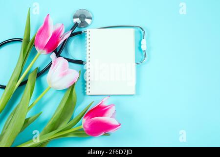 Tulipes roses et stéthoscope avec bloc-notes en spirale sur fond bleu. plan d'examen à plat vue du dessus, Banque D'Images