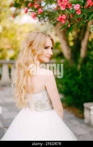 Une mariée blonde gracieuse se tient sous les branches d'un La lauriers-roses en fleurs près de l'ancienne église de Prcanj Banque D'Images