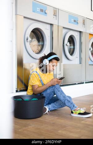 Jeune femme hispanique assise sur le sol à l'écoute de musique et à l'aide de son smartphone. Elle attend que ses vêtements soient lavés en libre-service l Banque D'Images