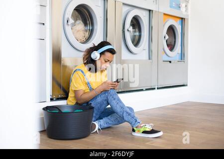 Jeune femme latina assise sur le sol pour écouter de la musique et utiliser son smartphone. Elle attend que ses vêtements soient lavés à un lau libre-service Banque D'Images