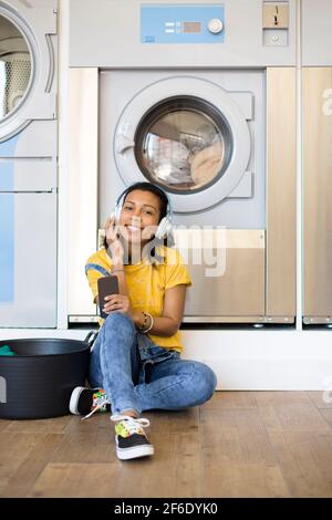 Jeune femme hispanique appréciant de la musique attendant que les vêtements soient lavés assis sur le sol à la blanchisserie libre-service. Espace pour le texte. Banque D'Images