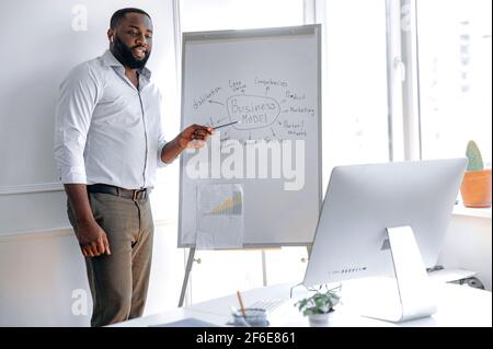 Formation à distance. Un mentor d'affaires afro-américain intelligent et fiable, conduit une conférence en ligne pour les étudiants, debout près d'un tableau de conférence, montrant des informations, regarde l'écran d'ordinateur Banque D'Images