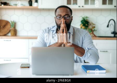 Entreprise concentrée afro-américain homme travaillant à la maison, assis au bureau, utilisant un ordinateur portable, regardant l'ordinateur portable de manière réfléchie, les mains près du visage, pensant à un plan d'affaires financier ou à une nouvelle start-up Banque D'Images