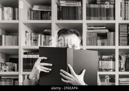 Portrait noir et blanc d'un homme qui a son face presque complètement couverte par un grand livre avec un bibliothèque carrée en arrière-plan Banque D'Images