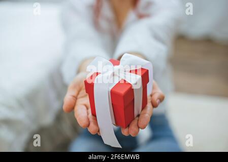 Boîte cadeau rouge avec ruban blanc dans les mains des femmes. Gros plan. Une fille dans une chemise blanche avec une boîte cadeau dans ses mains. Banque D'Images