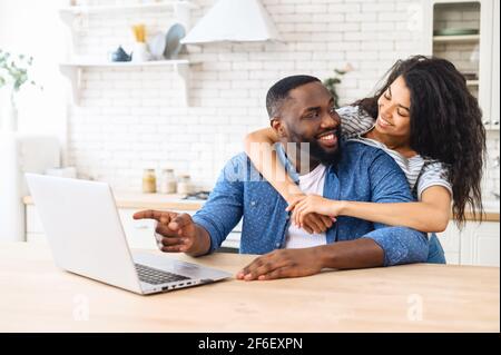 Un couple afro-américain heureux avec un ordinateur portable pour planifier des vacances ensemble, en utilisant l'application informatique pour réserver des hôtels ou une visite, homme souriant pointe le doigt sur l'écran, une femme l'embrasse et rit Banque D'Images