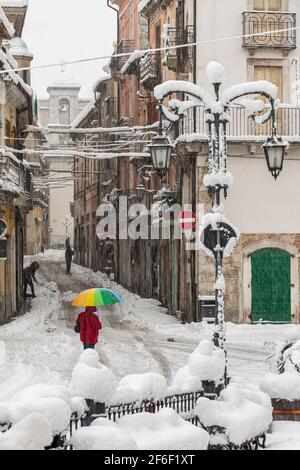 Rue principale d'une ville des Abruzzes sous une chute de neige avec une personne dans un manteau rouge et un parapluie de couleur. Pratola Peligna, Abruzzes Banque D'Images