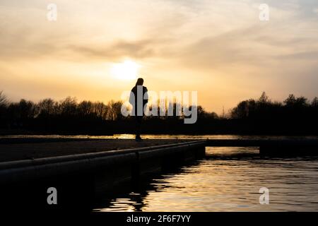Femme debout seule au coucher du soleil à l'extrémité de la jetée du lac jetée pensive et calme rivière ci-dessous. Soleil doré été Sin lumière se reflétant sur l'eau solitaire Banque D'Images