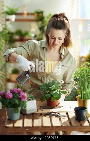 Maison verte. Jeune femme en gants de caoutchouc blanc à la maison moderne dans une plante d'arrosage de jour ensoleillé. Banque D'Images