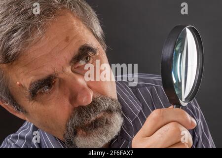 Un homme âgé aux cheveux gris avec une barbe examine soigneusement quelque chose à travers une loupe Banque D'Images