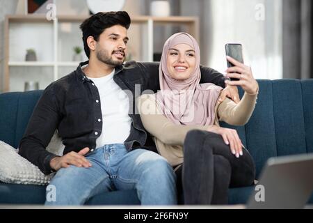 Vue rapprochée de l'avant d'un charmant jeune couple musulman gai assis sur un canapé à la maison, à l'aide d'un téléphone portable et faisant des selfies photo ensemble. Banque D'Images