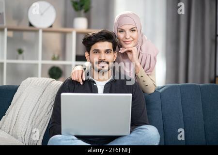 Joyeux jeune couple musulman décontracté travaillant sur un ordinateur portable à l'intérieur confortable de la maison. Beau homme assis sur un canapé avec un ordinateur portable, tandis que jolie femme en foulard l'embrassant de l'arrière. Banque D'Images