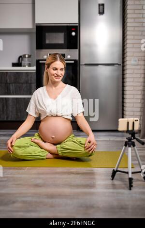 femme enceinte assise sur le sol dans une posture de yoga, avec des jambes croisées, enregistrant des vidéos sur un smartphone, menant un cours de yoga de traduction en ligne dans le salon Banque D'Images