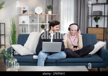 Apprentissage à distance et travail. Loisirs avec gadgets à la maison. Jeunes amis musulmans de 30 ans, hommes et femmes, assis sur un canapé dans un salon confortable et utilisant un ordinateur portable, un smartphone et des écouteurs. Banque D'Images