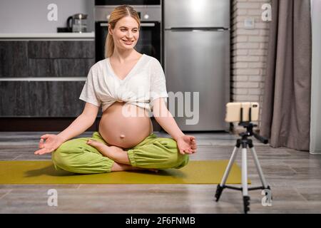 femme enceinte assise sur le sol dans une posture de yoga, avec des jambes croisées, enregistrant des vidéos sur un smartphone, diriger un cours de yoga de traduction en ligne dans le salon, à Banque D'Images