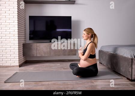 Agréable femme gravid méditant sur le sol à la maison, avec des jambes croisées, dans le Black Sportswear, Namaste, garder le calme. Vue latérale Banque D'Images