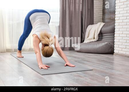 Femme blonde non reconnaissable faisant Adho Mukha Svanasana exercice à la maison seul, face vers le bas, sur le tapis de fitness, en vêtements sportifs. Jeune femme enga Banque D'Images