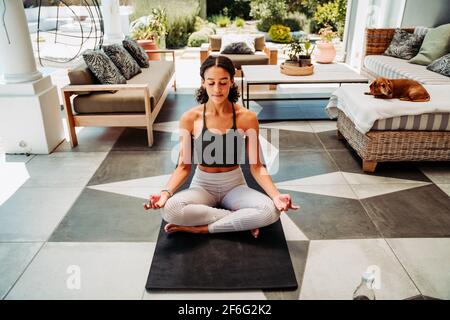 Femme de course mixte méditant assis en croix sur le tapis de gym à l'extérieur de la maison Banque D'Images