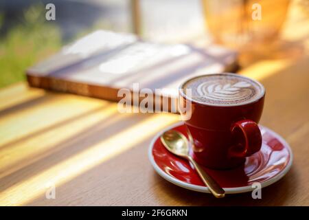 Gros plan chaud art Latte, cappuccino café dans une tasse rouge sur une table en bois dans le café-restaurant flou d'arrière-plan avec image bokeh. Banque D'Images