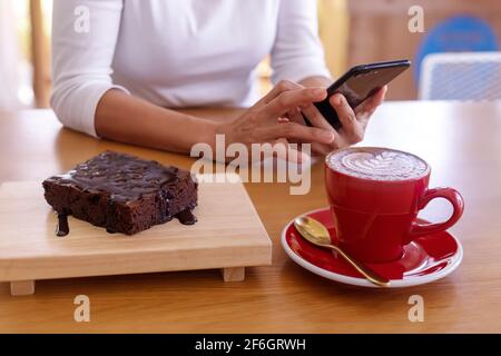 Femme mains tenant le smartphone mobile assis dans un café avec une tasse de café et des brownies dessert. Les femmes se détendent ou travaillent avec un téléphone portable dans un café Banque D'Images