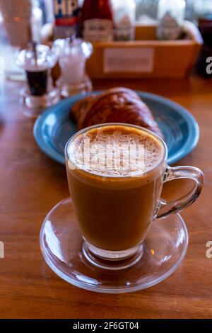 Gros plan sur le cappuccino chaud, le latte, le macchiato caramel sur la table avec un arrière-plan flou du café. Café cappuccino chaud sur table en bois avec croissant flou b Banque D'Images