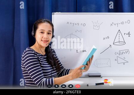 Femme d'école asiatique enseignant travaillant à la maison enseigner en ligne des mathématiques à l'étudiant étudiant étudiant étudiant à la maison. L'enseignant écrit sur le tableau blanc et parle sur l'interface micro-casque/oreillette Banque D'Images