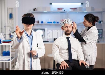 Neurologue médecin gestante portant le casque de réalité virtuelle et assistant de réglage du capteur de patient qui lit l'activité cérébrale , système nerveux. Neuroscientifique analysant le diagnostic. Banque D'Images