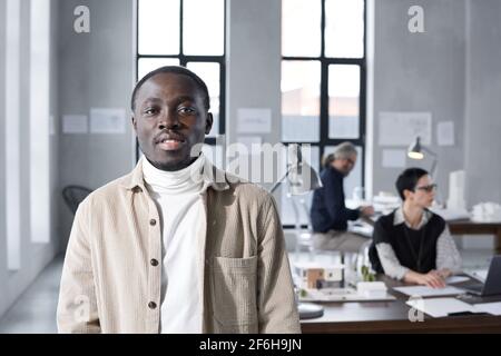 Portrait d'un jeune homme d'affaires africain regardant l'appareil photo debout au bureau moderne avec ses collègues en arrière-plan Banque D'Images