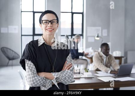 Portrait d'une femme d'affaires mûre réussie en lunettes souriant à l'appareil photo tout en étant au bureau avec ses collègues en arrière-plan Banque D'Images