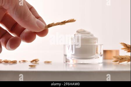 Crème hydratante pour le corps et le visage d'avoine dans un pot en verre avec pointes sur la table blanche et pointe de maintien à la main avec du blanc arrière-plan isolé Banque D'Images