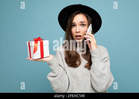 Plan de la jeune femme brunette assez triste choquée isolée le mur sur fond bleu porte un élégant chapeau noir et un chandail gris boîte cadeau en attente en conversation Banque D'Images