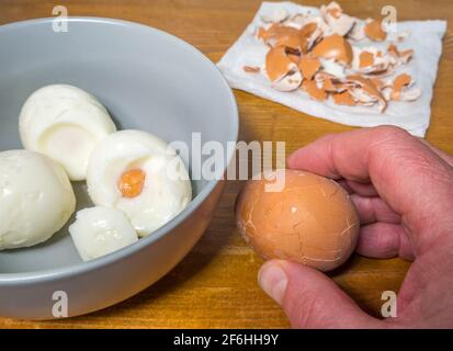 Gros plan de la main d’un homme qui fend un œuf dur sur un plan de travail ou un comptoir en pin, à côté de quelques coquilles retirées sur une serviette et d’un bol d’œufs pelés. Banque D'Images