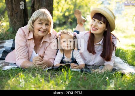 Grand-mère, mère et petite fille appréciant des vacances ensoleillées dans le jardin ensemble à l'extérieur, allongé sur l'herbe verte sur une couverture et souriant à l'appareil photo. Loisirs famille mode de vie, bonheur et moments. Banque D'Images