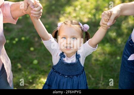 Top vue rapprochée de rire adorable 3 ans enfant fille en robe Jean, s'amuser et profiter de sa promenade à l'extérieur dans le parc, tenant les mains de sa mère et de la grandmothe. Promenade en famille dans le jardin. Banque D'Images