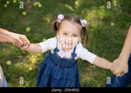 Top vue rapprochée de rire adorable 3 ans enfant fille en robe Jean, s'amuser et profiter de sa promenade à l'extérieur dans le parc, tenant les mains de sa mère et de la grandmothe. Promenade en famille dans le jardin. Banque D'Images