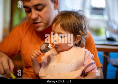 un adorable tout-petit aime manger de la glace à la popsicle sur les genoux de ses parents dans l'intérieur de la maison Banque D'Images