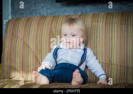 Un petit garçon, un enfant, s'assoit sur le canapé et attend ses parents. Dans une belle tenue, une chemise blanche et un pantalon avec bretelles. Banque D'Images