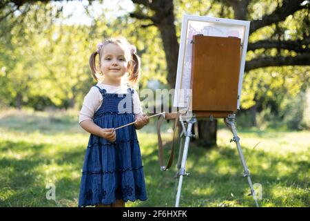 Gros plan portrait extérieur de la adorable petite fille en robe denim, posant à l'appareil photo avec la brosse dans les mains, tout en se tenant près d'un chevalet avec toile, peindre une photo dans le parc vert d'été. Banque D'Images