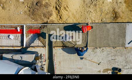 Au-dessus de la vue de dessus, les ouvriers du bâtiment utilisent le râteau pour l'épandage et la latte métallique pour niveler le béton frais dans la tranchée carrée après le versement Banque D'Images