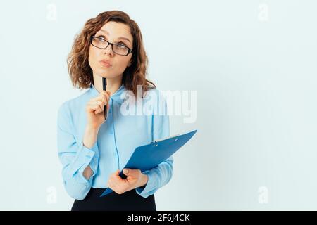 Jeune femme d'affaires pensive portant des lunettes et une chemise bleue tenant un stylo et un bloc-notes debout sur fond blanc studio avec espace pour le texte. Banque D'Images