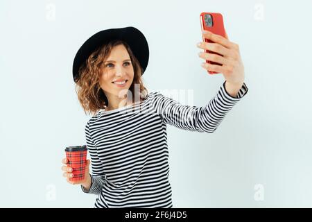 Une jeune femme souriante portant un chapeau fedora et une chemise rayée boit du café et fait du selfie à l'aide de son smartphone, fond blanc. Banque D'Images