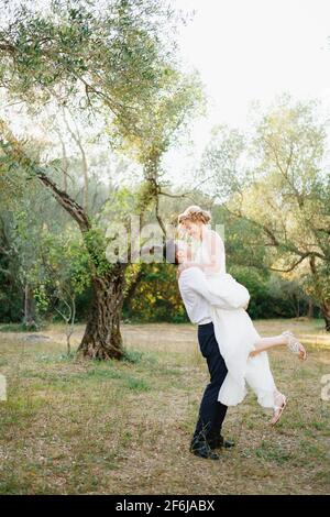 Le marié entoure la mariée dans ses bras parmi les arbres de l'oliveraie, la mariée sourit Banque D'Images