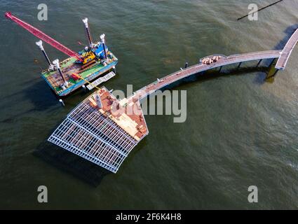 22 mars 2021, Mecklembourg-Poméranie occidentale, Koserow: Un ponton flottant avec une grue se trouve à côté de la nouvelle jetée en construction sur l'île d'Usedom. En raison du remplacement nécessaire de certains bois tropicaux posés comme planches de course et de la galvanisation renouvelée des segments de rampe, le pont ne sera maintenant prêt pour le festival de pont qu'au début de juillet 2021. La jetée s'étend sur près de 300 mètres dans la mer Baltique, et la forme de vague incurvée de la jetée droite sur d'autres ponts est frappante. La nouvelle construction coûtera plus de cinq millions d'euros. (Photo prise avec un drone) photo: Jens Büt Banque D'Images